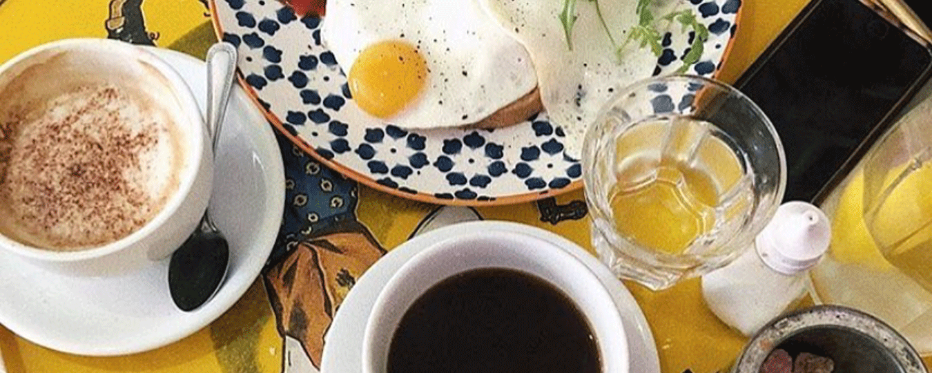William Cafe image of two coffees with a plate with fried eggs on a yellow table